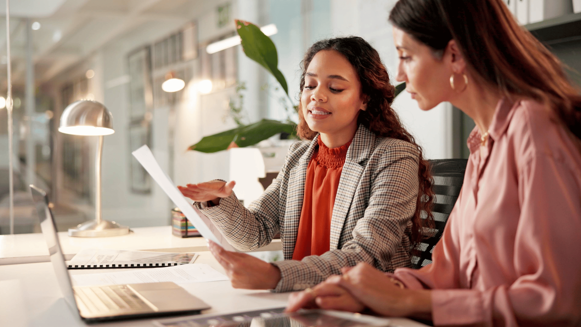 Two woman dressed in business casual attire site a table reviewing a document in front of a laptop.