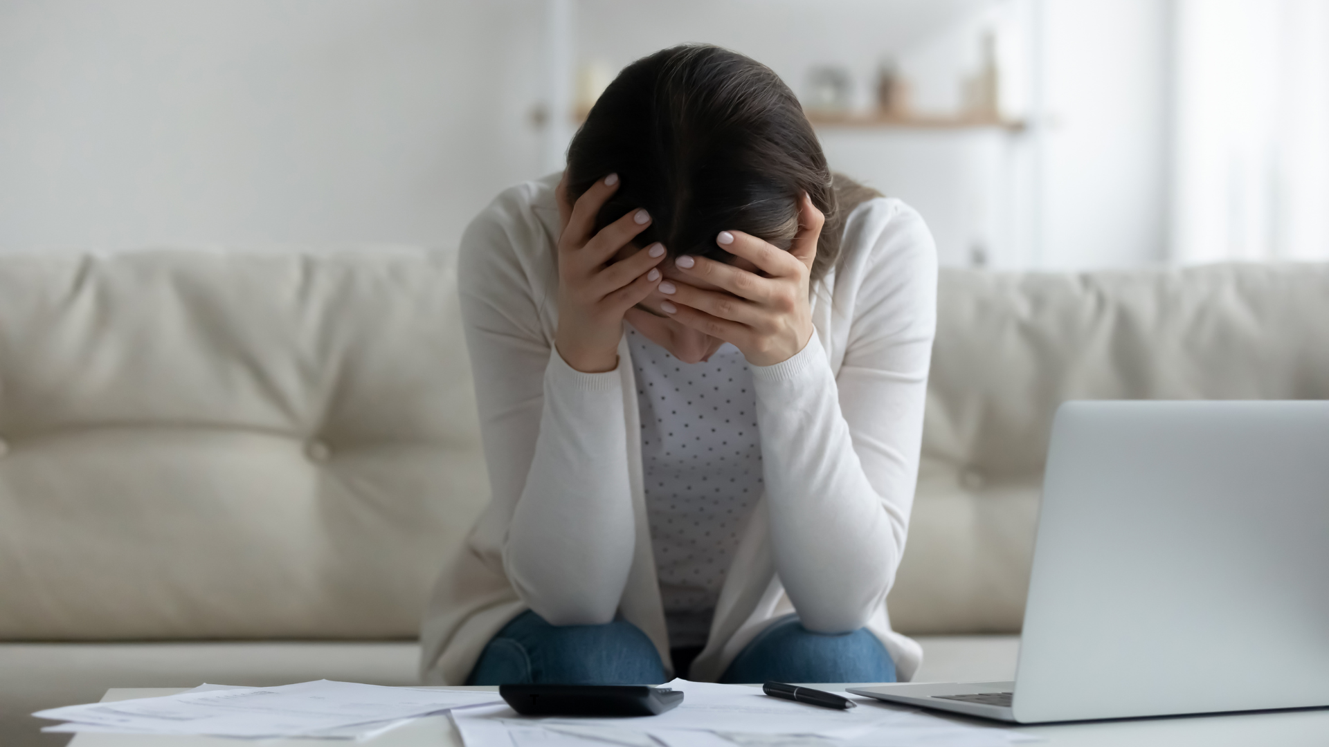 Woman holding her head in her hands over a table covered in papers and a laptop